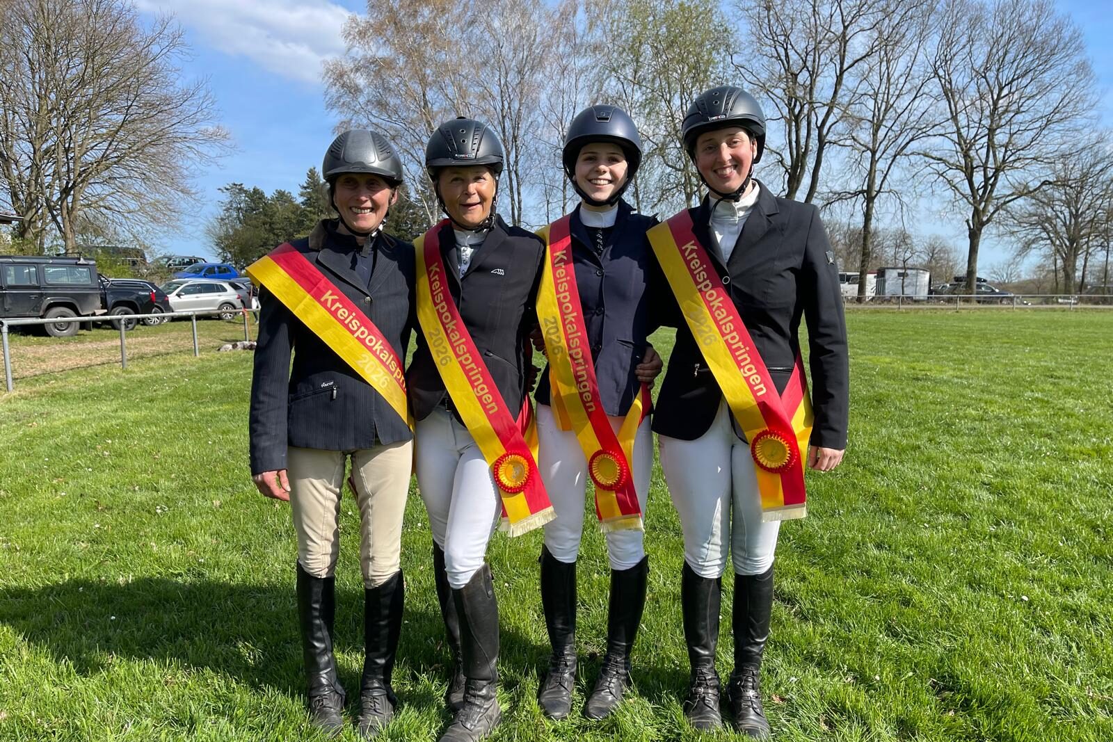 Sieger_Kreispokalspringen_2026 Vier Reiterinnen in schwarzen Jacken mit schwarz-rot-goldenen Schärpen beim Kreispokalspringen 2026 lächeln gemeinsam auf einer Wiese